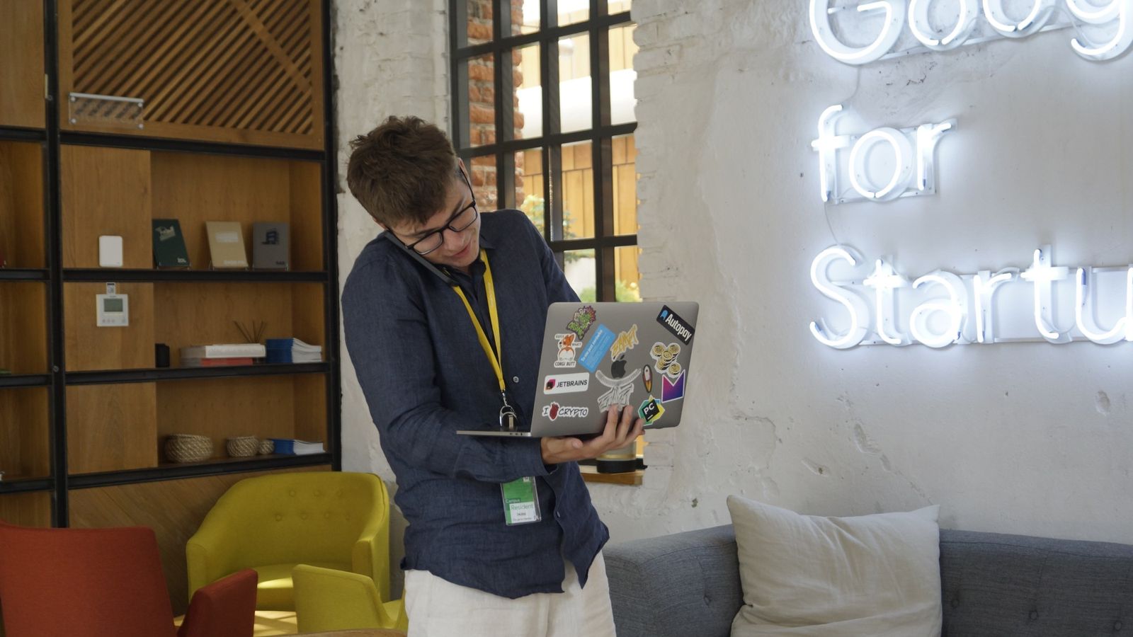 Mikita on a phone call, holding an open laptop, next to the Google for Startups neon sign.