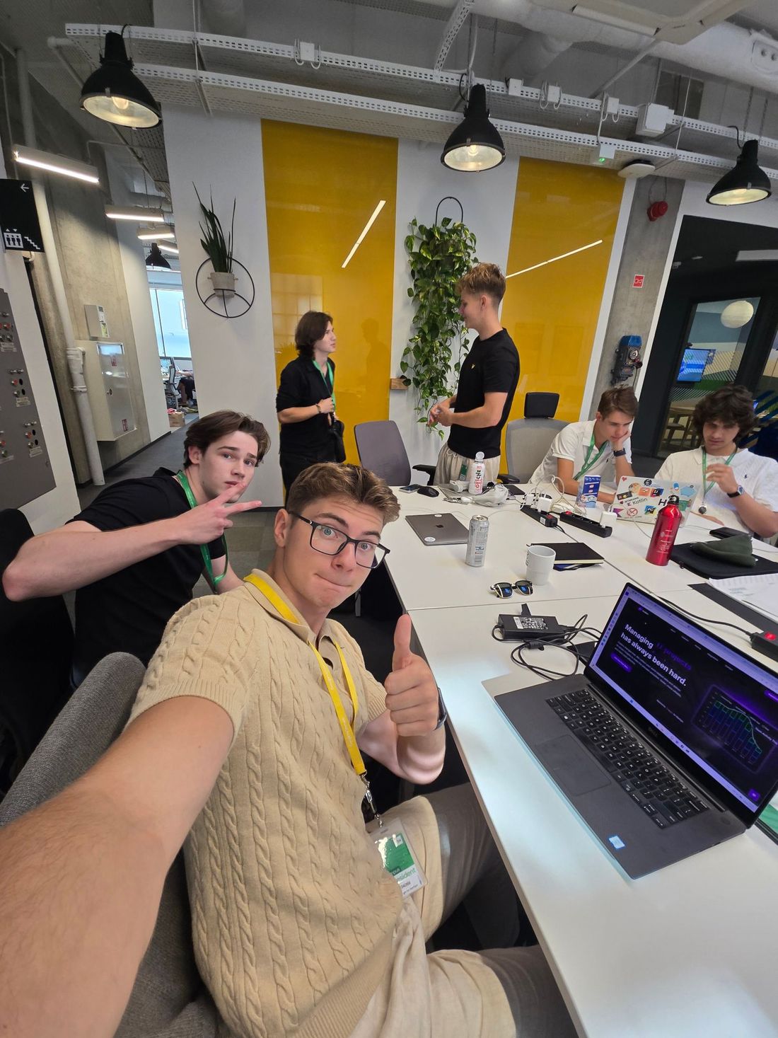 Selfie of Mikita giving a thumbs up at a shared desk, with teammates working behind him and the Codaro landing page on his laptop.