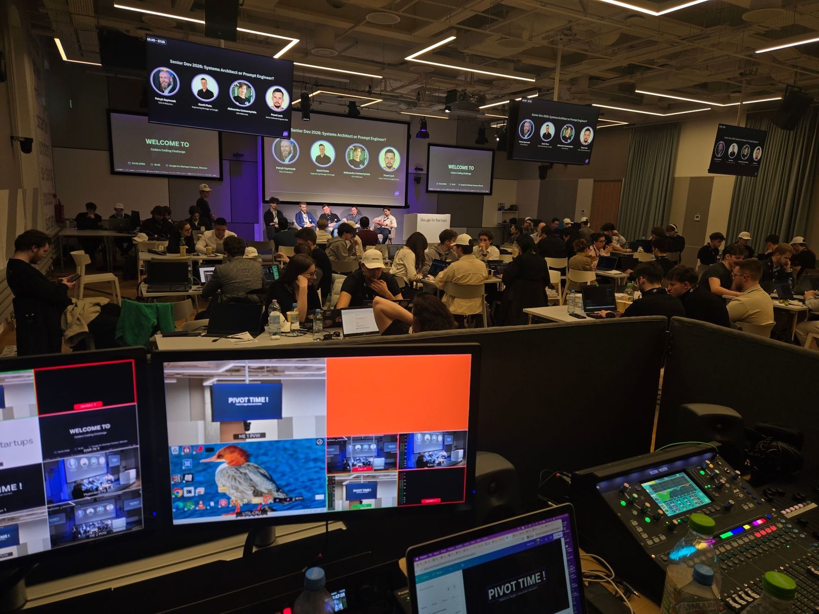 View from the production desk over a full hall of hackathon participants at laptops, with a four-person panel on stage and Welcome to Codaro Coding Challenge on the screens.