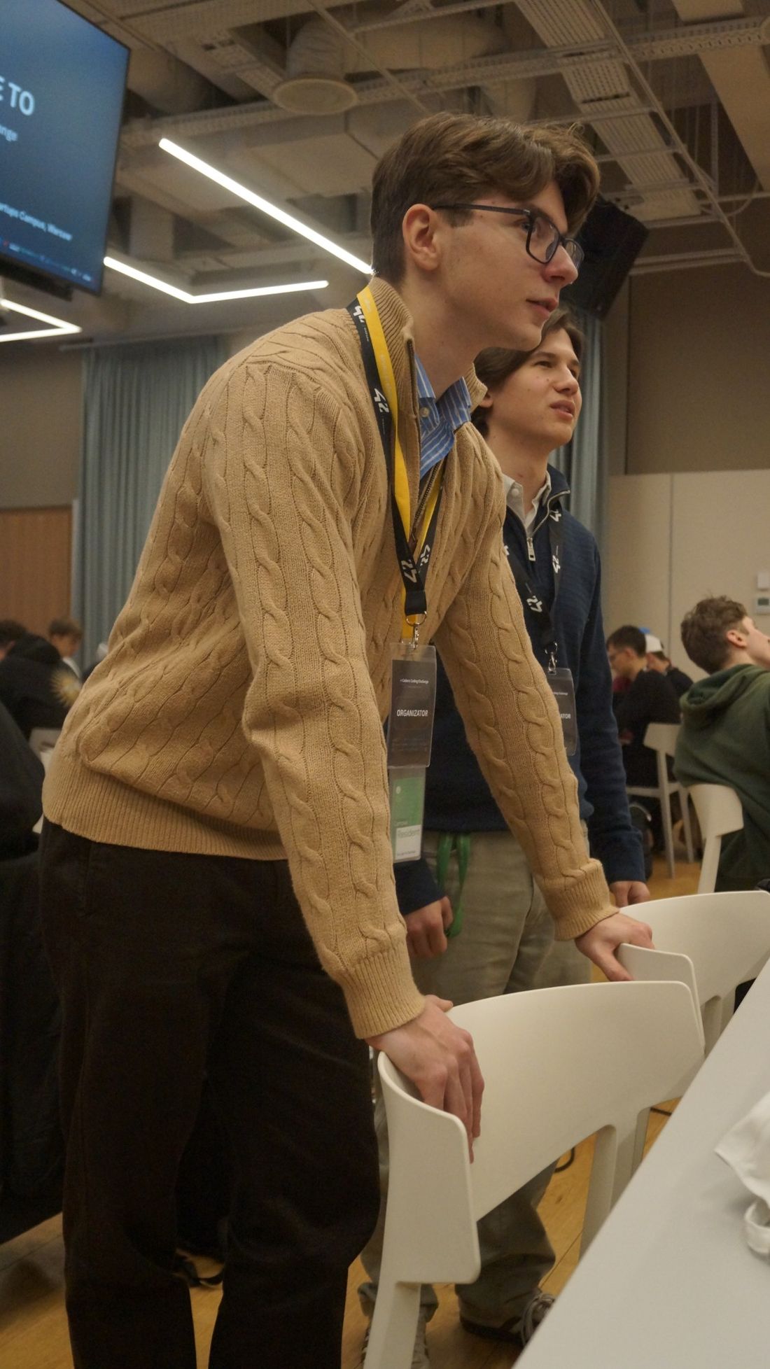 Mikita with an organiser badge, leaning on a chair and watching the hackathon, with a teammate beside him.