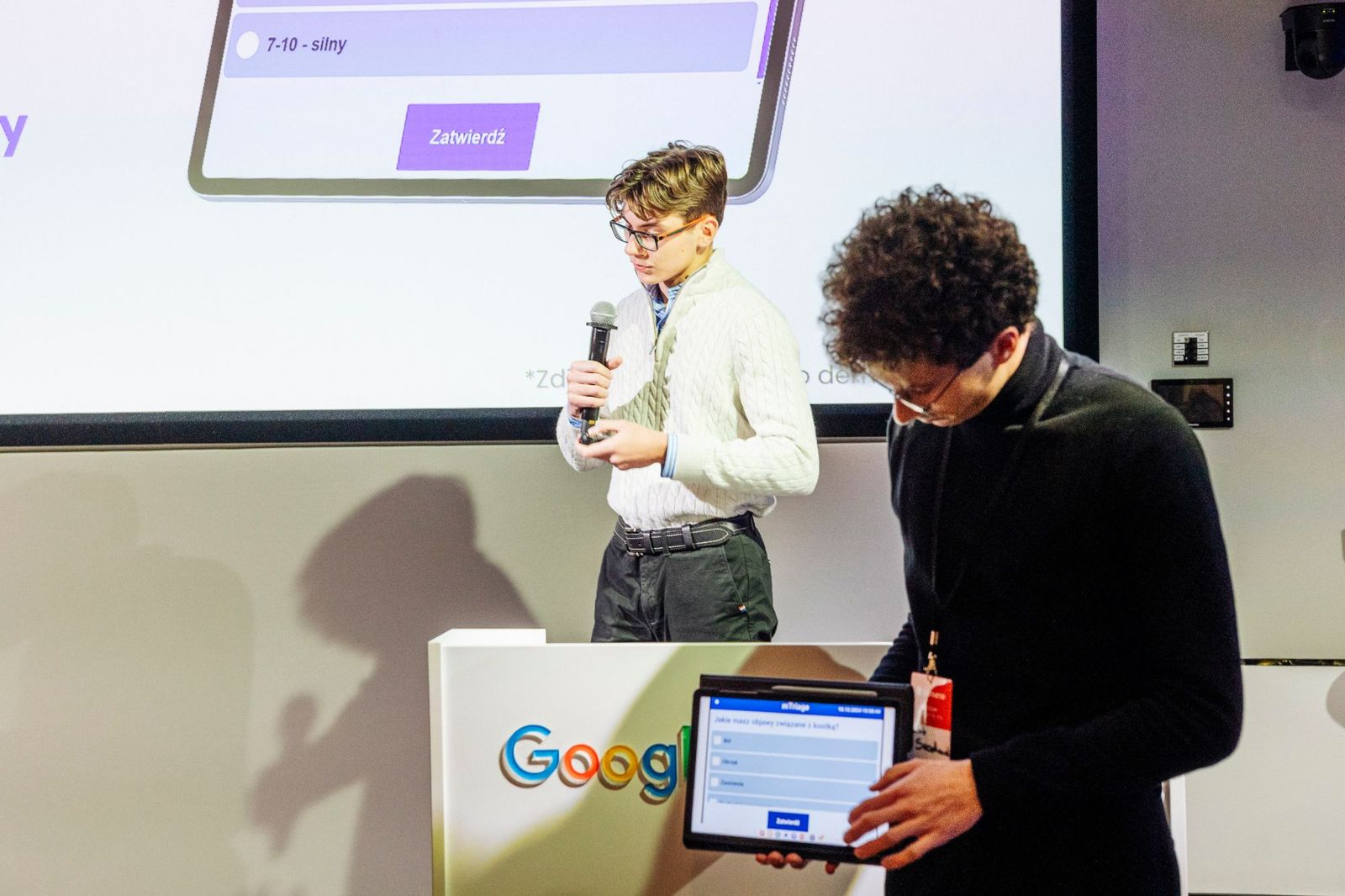 Mikita presenting on stage with a microphone while another person holds a tablet showing the first version of mTriage, a patient symptom questionnaire.
