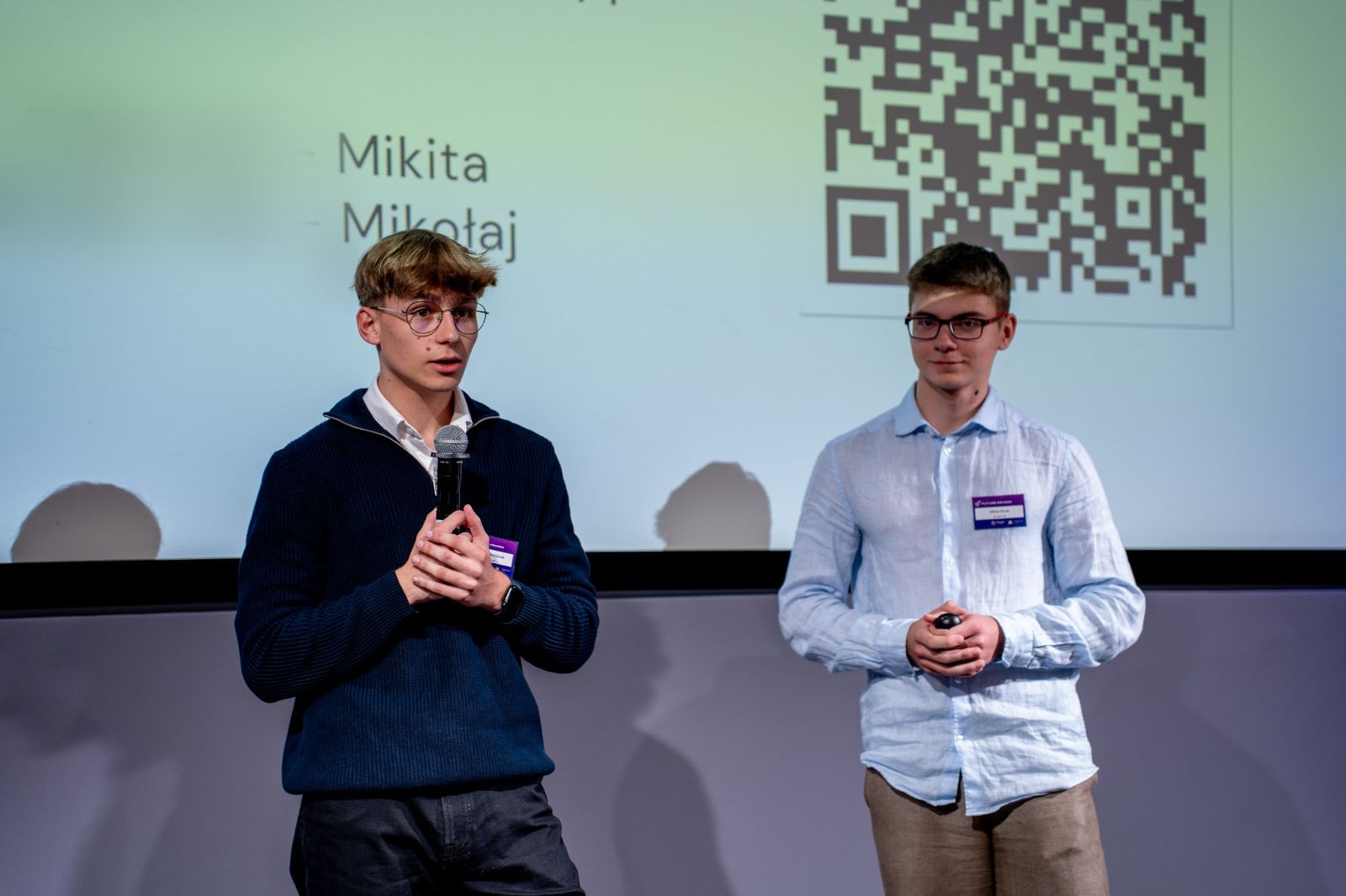 Mikołaj speaking into a microphone and Mikita standing next to him on stage, in front of the closing slide of their pitch.