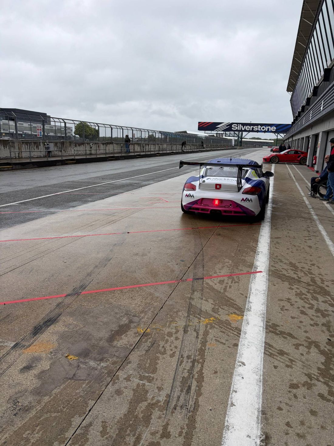 A race car in the pit lane at Silverstone on a wet day, with a red Porsche behind it.
